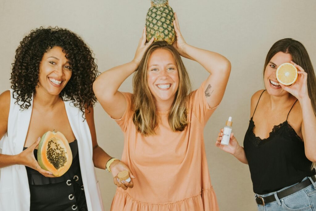 Three women smiling while holding pineapples, papaya, and oranges symbolizing joy and health.